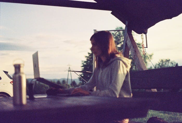 Photo of the author — a woman with brown hair a bit past shoulder-length wearing a hoodie, sitting by a wooden table outside, working on her laptop. Trees and hills are visible in the distance.